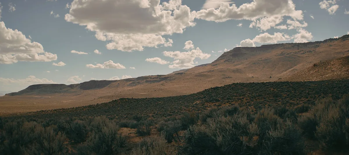 Sagebrush in the foreground below long, flat-topped desert ridges under scattered white clouds.